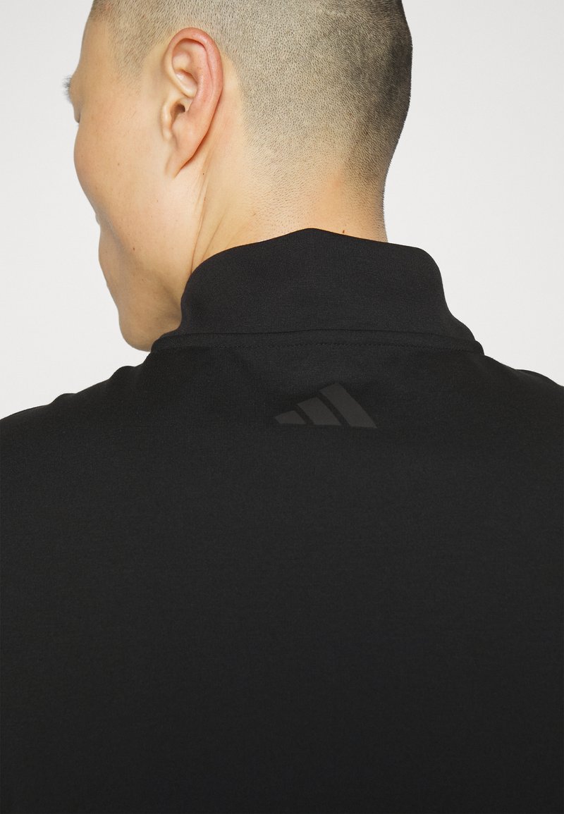 Zwarte polo met een hoge kraag, gemaakt van een gladde stof. Heeft een subtiele zwarte Adidas-logo op de rug.