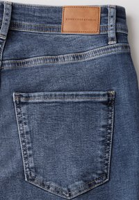 Jeans en denim avec un lavage bleu, dotés d'une poche arrière centrale avec des coutures contrastées et d'une étiquette en cuir brun portant la mention "STREET ONE STUDIO."