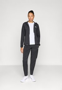 Μαύρο αθλητικό μπουφάν με κουκούλα, συνδυασμένο με αντίστοιχο παντελόνι. Διαθέτει ελαστικά μανίκια, φερμουάρ μπροστά και λευκά λογότυπα. Λευκά αθλητικά παπούτσια.