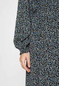 Main reposant près de la taille, portant une robe noire avec de petits motifs floraux bleus et blancs et une manche avec un poignet élastique.