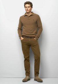 Junger Mann mit lockigem Haar, der einen braunen Strickpullover, passende braune Hosen, einen mehrfarbigen Gürtel und beiges Wildlederstiefel trägt, steht mit den Händen in den Taschen.