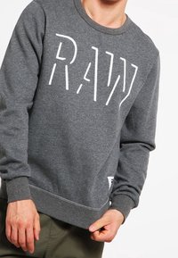 Sudadera gris con cuello redondo, que presenta un logotipo blanco de "RAW". Fabricada con material suave y con puños y dobladillo acanalados.