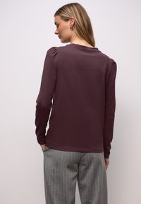 Langärmeliges Hemd aus dunkelviolettem Stoff mit glatter Textur, versehen mit gepufften Schulterdetails und rundem Halsausschnitt. Kombiniert mit grauer Hose mit Nadelstreifen.