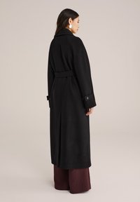 Schwarzer, langer Mantel aus weichem Stoff, mit einem taillierten Gürtel, großen Revers und geknöpften Manschetten, kombiniert mit weiten, burgunderroten Hosen.