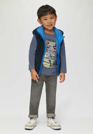 Blauwe fleecevest over een grijs langetermijnshirt met een grafische print, gecombineerd met grijze jeans en witte sneakers. Geen opvallende patronen.