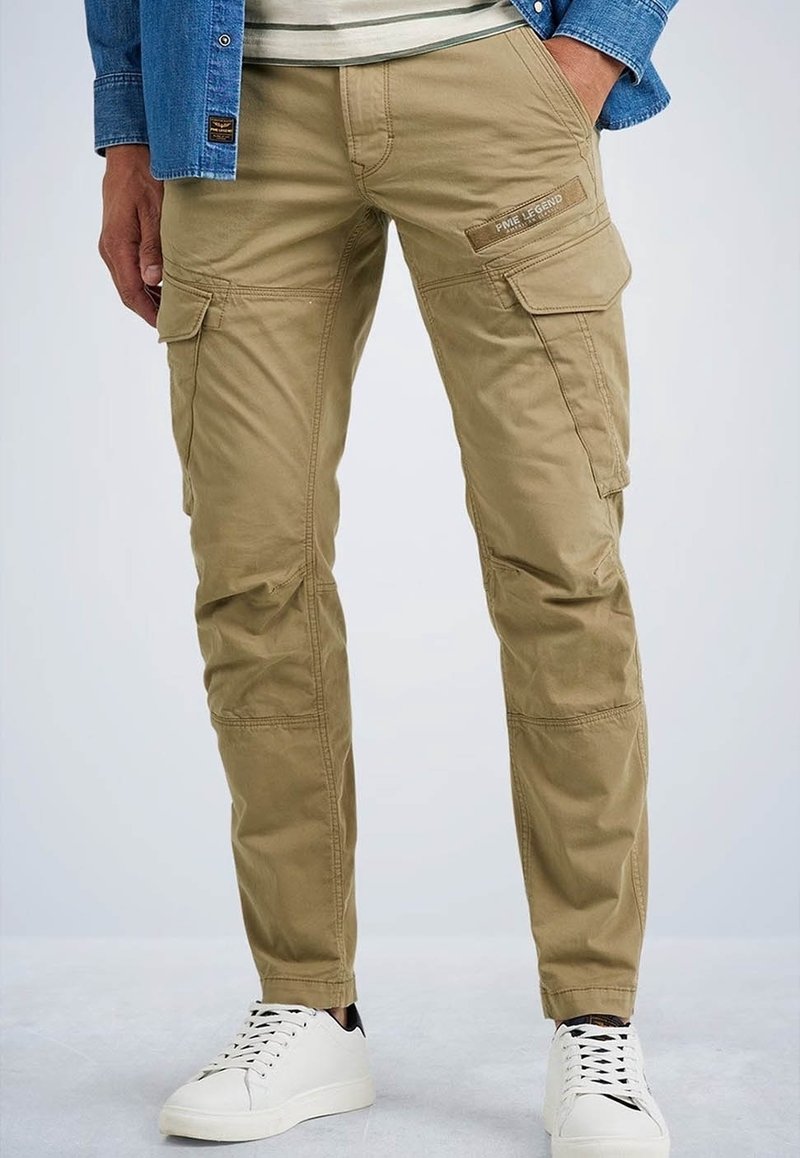 Homme portant un pantalon cargo beige avec des poches à rabat et des baskets blanches, debout avec une main dans la poche sur un fond uni.