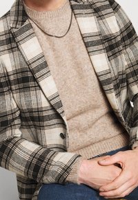 Gepunkteter Blazer in schwarz-weißer Karoprint über einem beigen Strickpullover. Enthält eine Kettenhalskette. Die Hände sind gefaltet auf einer dunklen Jeans.