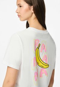 Valkoinen puuvillapaita, jossa on keltainen banaanigrafiikka ja vaaleanpunainen teksti "BANANA" selässä. Lyhyet hihat ja klassinen pyöreä kaula-aukko.