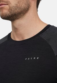Chemise noire à manches courtes avec accents rayés aux épaules; présente une texture lisse et un logo subtil "FALKE" sur la poitrine.