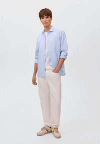 Camicia button-up azzurro chiaro sopra una t-shirt bianca, abbinata a pantaloni crema e sneakers beige con dettagli verdi.
