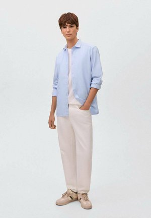 Chemise à boutons bleu clair sur un t-shirt blanc, associée à un pantalon crème et des baskets beiges avec des accents verts.
