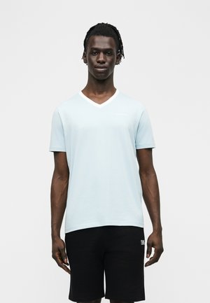 Jaunas vyras su pintais plaukais, vilkintis šviesiai mėlyną V formos kaklo marškinėlį ir juodus šortus, stovintis ant paprasto balto fono.