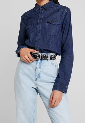 Henkilö, joka pukeutunut tummansiniseen farkkupaitaan, joka on sullottu vaaleansinisten korkeavyötäröisten farkkujen sisään, mustalla niitityllä vyöllä.