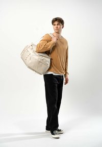 Sac de sport beige avec une surface texturée, comportant plusieurs compartiments et une large sangle de portage. Porté par une personne dans un pull marron.