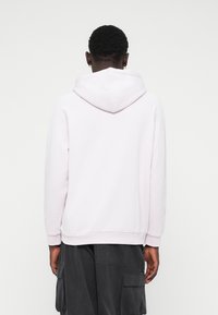 En person med kort, lockigt hår som bär en ljusrosa hoodie och mörka cargobyxor, vänd bort från kameran mot en enkel vit bakgrund.