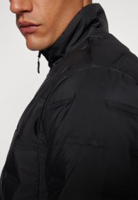Black padded jacket with a high collar, featuring textured accents, sleek design, and horizontal stitching detail along the sleeves.