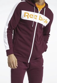 Felpa con zip color bordeaux e maniche bianche, con un grande logo giallo "Reebok". Presenta una tasca frontale e cappuccio regolabile. Indossata con pantaloni coordinati.