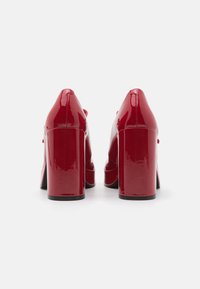 Scarpe con tacco alto in vernice rossa con tacco spesso viste da dietro su uno sfondo bianco semplice.