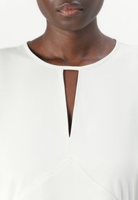 Lys grå kjole med en nøglehul-neckline og vinklede sømdetaljer. Fremstillet af et glat stof med en subtil glans.