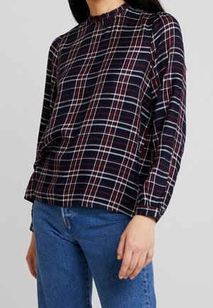 Woman wearing a navy, red, and white plaid long-sleeve top with ruffled neckline and blue jeans, standing against a plain background.