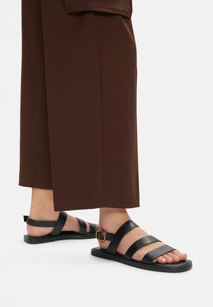Sandali neri in pelle con più cinturini e dettaglio con fibbia. Suola piatta, design minimalista, abbinati a pantaloni larghi marroni.