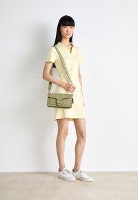 Kobieta w jasnożółtej sukience z krótkim rękawem, w białych sneakersach i trzymająca oliwkową torbę na ramię, stojąca przy białej ścianie.