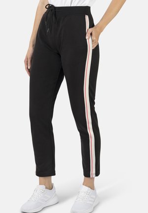 Schwarze Jogginghosen mit weißen und roten Seitenstreifen, getragen mit weißen Sneakern, Hände in den Taschen, dargestellt von der Taille bis zu den Füßen.