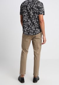 Lyhythihainen musta kukkapaita, jossa on valkoisia ja beigejä kukkia, yhdistettynä ruskeisiin kapeisiin housuihin ja mustiin kenkiin. Näkymä takaa.