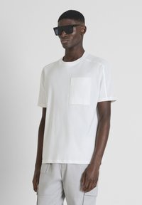 T-shirt bianco a maniche corte con taschino sul petto, pannelli laterali strutturati e colletto rotondo. Indossato con pantaloni cargo grigi e occhiali da sole neri.