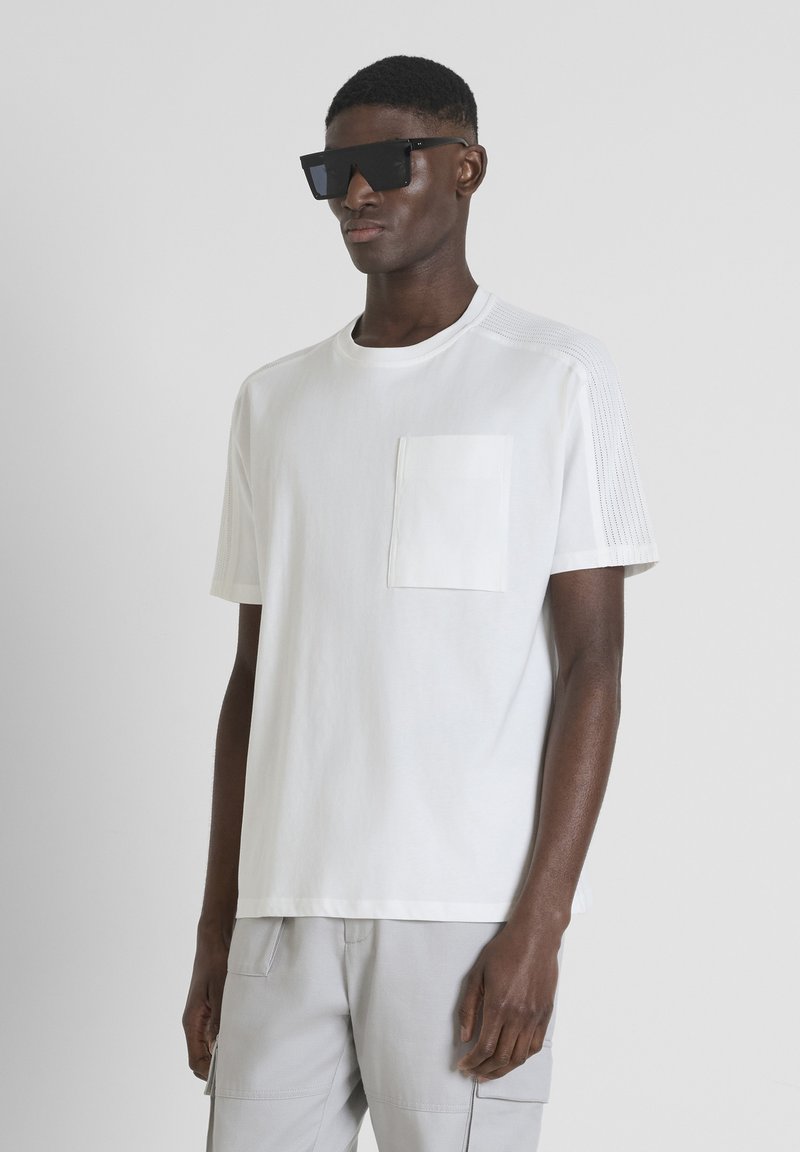 T-shirt bianco a maniche corte con taschino sul petto, pannelli laterali strutturati e colletto rotondo. Indossato con pantaloni cargo grigi e occhiali da sole neri.