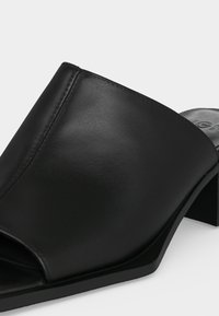 Mule en cuir noir à bout ouvert avec empeigne large, talon bloc et finition mate lisse, présentée sur un fond clair uni.
