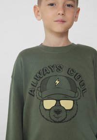 Olivově zelená mikina s grafikou medvěda v brýlích a s kšiltovkou. Nápis "VŽDY COOL" je černě nad designem. Měkká tkanina.