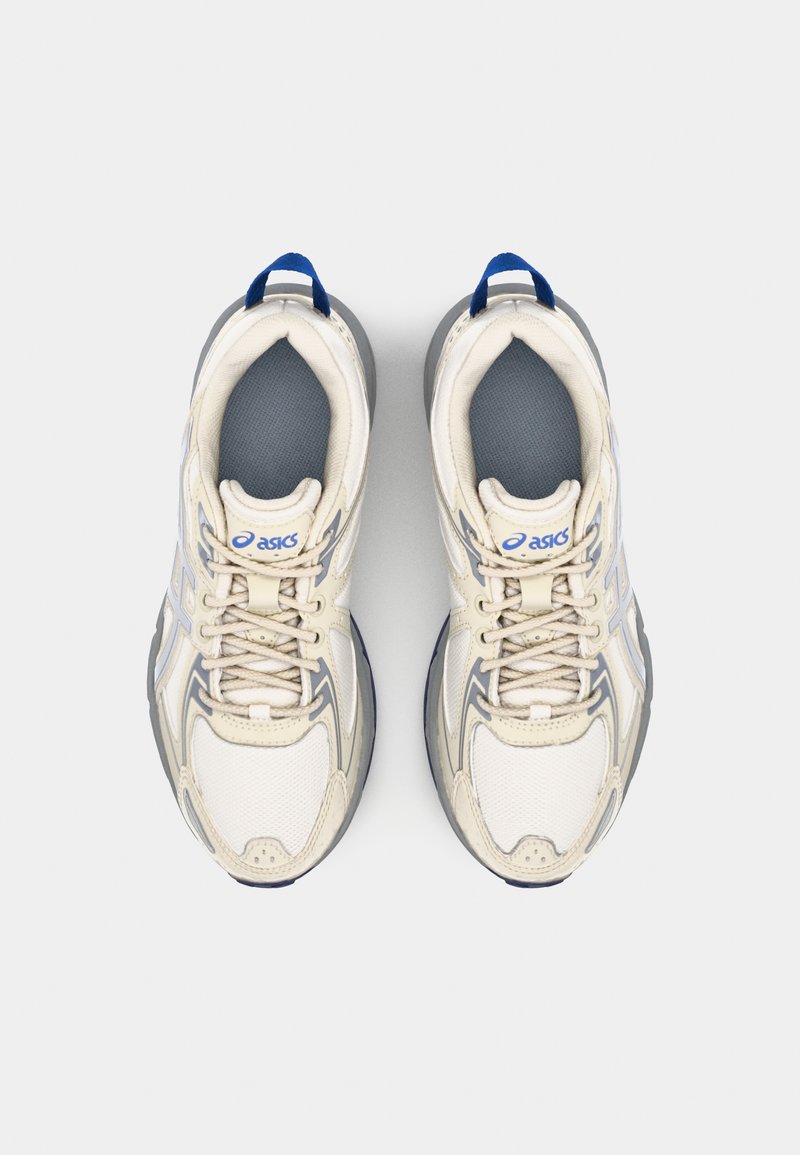 Zapatillas deportivas beige con acentos grises, parte superior de malla, diseño con cordones, cuello acolchado y etiqueta azul en el talón. Situadas en posición plana.