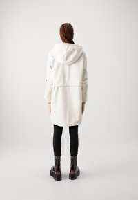 Hvid hættejakke med indsnøret talje, der har lange ærmer og en glat tekstur. Kombineret med sorte teksturerede leggings og blanke ankelstøvler.