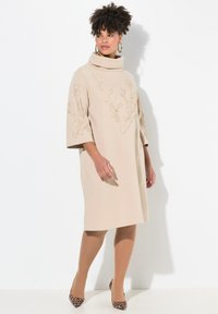 Beiges Kleid mit hohem Kragen, versehen mit floraler Stickerei und Ellenbogen-langen Ärmeln. Kombiniert mit hautfarbenen Strumpfhosen und Schuhen mit Leopardenmuster.