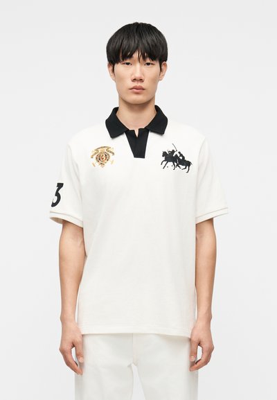 Joven de pie mirando al frente con camisa tipo polo blanca con cuello negro, logotipo de jugador de polo a caballo en el lado izquierdo del pecho, número 3 en la manga derecha.