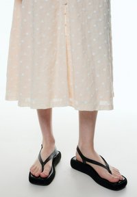 Schwarze Gummiflip-Flops mit Plattformsohle und V-förmigen Riemen, kombiniert mit einem hellpfirsichfarbenen Kleid, das weiße Punkten hat.