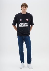 T-shirt nera dei Giants NFL con testo bianco, presenta un'etichetta con logo, abbinata a jeans blu e scarpe bianche, indossata da un modello su uno sfondo bianco.