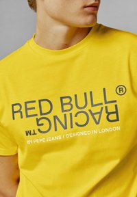 Žlté tričko s veľkými sivými nápismi "RED BULL RACING" a "BY PEPE JEANS / DESIGNED IN LONDON" potlačenými na prednej strane.