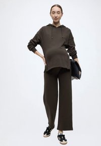 Σκούρο πράσινο, υπερμεγέθες φούτερ με κουκούλα και τσέπη καγκουρό, συνδυασμένο με αντίστοιχο παντελόνι φαρδιάς γραμμής. Το μοντέλο φοράει μαύρα αθλητικά παπούτσια και κρατά μια μαύρη τσάντα.