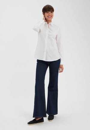 Chemise blanche à boutons, manches longues ; associée à un jean flare bleu foncé. Le mannequin prend une pose détendue, avec des espadrilles noires décontractées.