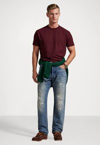 Μπλούζα μπορντό με στρογγυλή λαιμόκοψη, πράσινο πουλόβερ δεμένο στη μέση, φθαρμένα ανοιχτό μπλε τζιν, καφέ σουέντ παπούτσια. Απλή καθημερινή εμφάνιση.