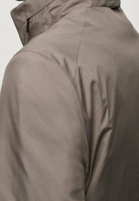 Leichte, braune Jacke mit hohem Kragen, die eine glatte Textur aufweist und eine externe Reißverschlusstasche an der Seite hat.