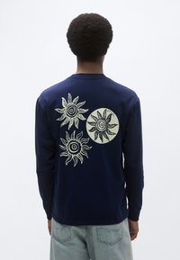 Μπλε σκούρο πουκάμισο με μακριά μανίκια, με τρεις απεικονίσεις του ήλιου σε ανοιχτό χρώμα στην πλάτη, με έναν στρογγυλό ήλιο στο κέντρο.