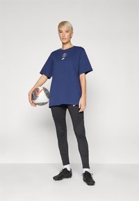 Mørkeblå oversized t-skjorte med logo, svarte tettsittende bukser med teksturerte mønstre, og svarte fotballsko, holder en fotball.