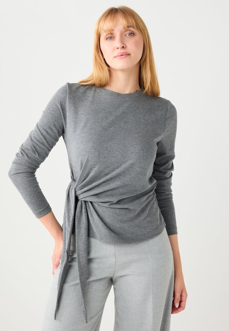 Haut gris à manches longues avec un détail nœud torsadé à la taille, en tissu doux. Associé à un pantalon gris clair pour un look coordonné.