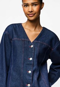 Denim jakna s V-izrezom, srebrnim gumbima s crvenim šavovima, tamnoplave boje i opuštenim krojem s širokim rukavima.