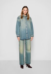 Camicia di denim con una sfumatura che va dal blu chiaro al giallo pallido, con bottoni davanti e maniche lunghe; abbinata a jeans a taglio largo e tacchi marroni.