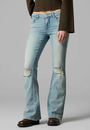 Kvinne iført lyseblå vide jeans med revne knær, brun cropped genser og mørkebrune sko som står mot en grå bakgrunn.