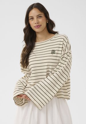 Mladá žena s dlhými tmavými vlasmi, ktorá má na sebe krémovú pruhovanú blúzku s dlhým rukávom a bielu sukňu, stojaca pred jednoduchým pozadím.